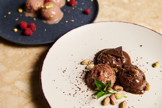 Selective focus on refreshing raw vegan frozen balls of chocolate pistachio ice cream, garnished with lemon basil on white plate, with partial view of raspberry sorbet on blue plate on the background