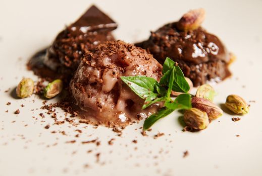 Selective focus on scoops of chocolate ice cream, topped with pistachio and lemon basil leaves, melting on a white plate. Food still life with copy space for advertising text. Refresshing summer snack