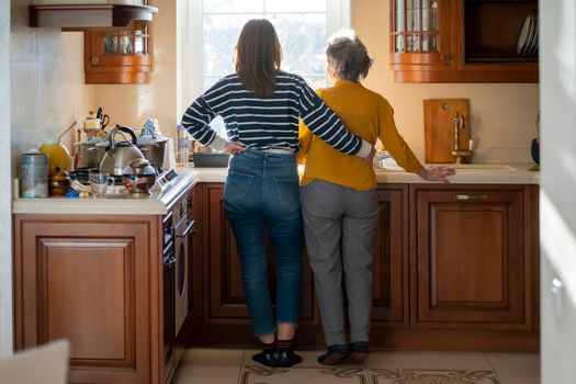The daughter hugs her elderly mother, the granddaughter and her grandmother enjoy life, look out the window at a beautiful view from their cozy home, spend time preparing together in the kitchen.