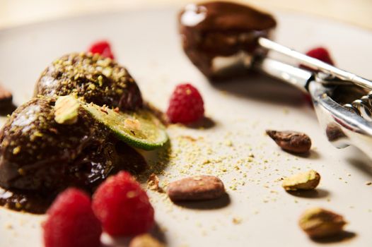 Flat lay of frozen chocolate ice cream balls, garnished with pistachios, cocoa beans, raspberries and slice of juicy lime. Blur metal scoop filled with chocolate sorbet on a white plate on background