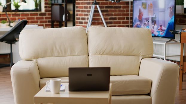 Interior of modern apartment with coffee table having laptop on it. Empty living room with nobody in it and with comfortable sofa and desk having portable computer on top of it.