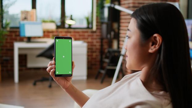 Cheerful person sitting on couch with mobile cellphone having chroma key mockup display background. Happy smiling heartily woman having smartphone with green screen display while sitting at home on sofa.