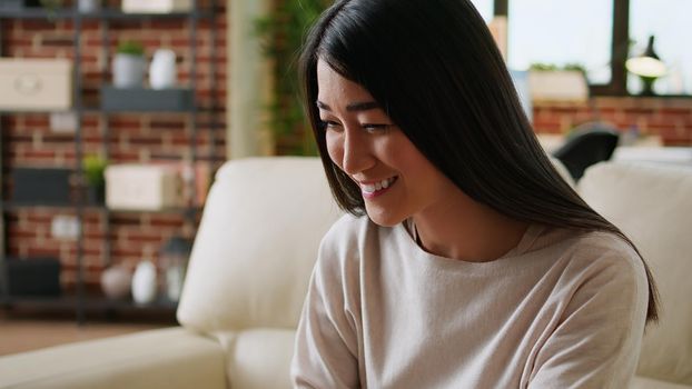 Excited young woman sitting on sofa at home while working remotely on laptop. Smiling heartily young adult asian person doing remote work on portable computer inside apartment.