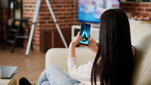 Childish asian person enjoying mobile gaming while sitting on sofa in modern apartment. Playful woman working remotely while playing space shooter game on smartphone at home.