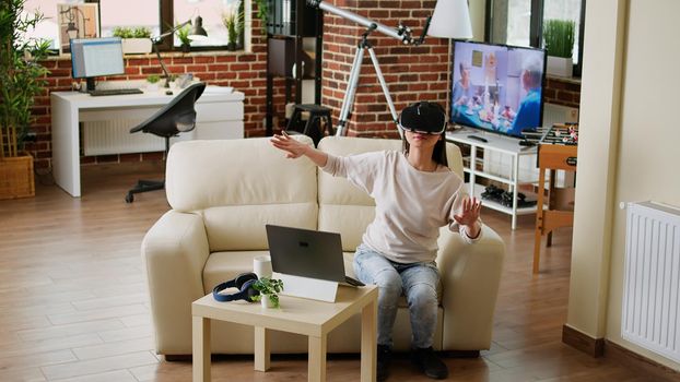 Young adult person wearing futuristic virtual reality headset enjoying cyberspace while sitting on sofa at home. Playful woman with VR goggles playing on metaverse inside living room.