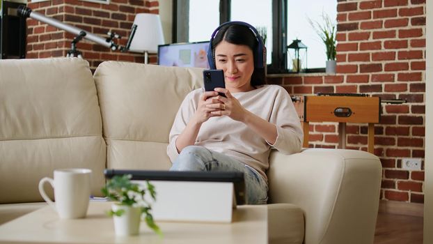 Happy woman browsing social media on smartphone while wearing headphones and working remotely from home. Cheerful smiling heartily person chatting on mobile phone while doing remote work.