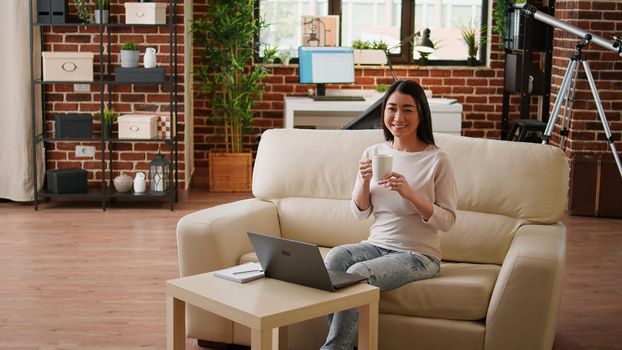 Beautiful student sitting on couch inside modern and cozy apartment while smiling heartily at camera. Attractive young adult person sitting in house while working remotely.