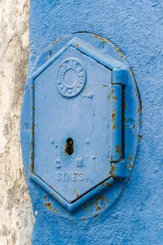 Old rusty blue painted iron lid of a valve box of the SERVICO DE AGUA PUBLIC WATER SUPPLY on the chipped white - blue painted wall of a house in the Old Town area, Portugal