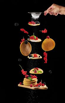 Levitating round pancakes, various fruits on a black background. Hand sprinkled with powdered sugar delicious traditional breakfast