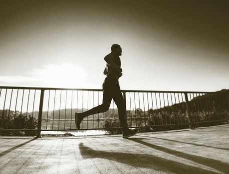 Tall man with sunglasses, red baseball cap and blue black sportswear is running and exercising on sea bridge. Sport active man.