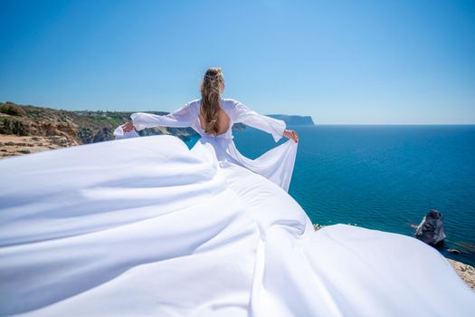 Blonde with long hair on a sunny seashore in a white flowing dress, rear view, silk fabric waving in the wind. Against the backdrop of the blue sky and mountains on the seashore