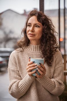 A middle-aged woman in a beige sweater with a blue mug in her hands is in a street cafe on the veranda.