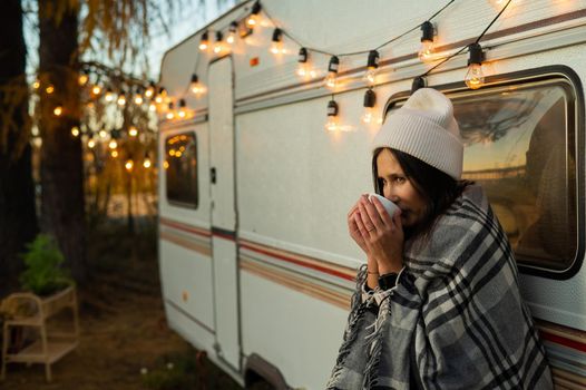 Caucasian woman in a knitted hat wrapped in a plaid and drinks a warming drink outdoors. Travel in a motor home in the fall