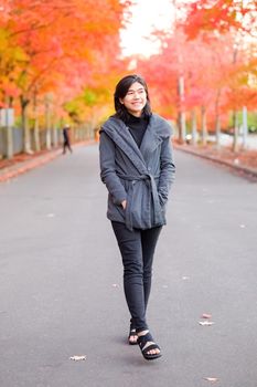 Smiling biracial teen girl or young adult female in gray jacket walking along road enjoying colorful autumn leaves