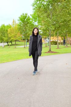 Smiling biracial teen girl or young adult female in gray jacket walking along road enjoying colorful autumn leaves