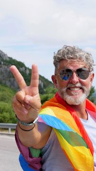 Portrait of a gray-haired senior elderly Caucasian man bisexuality with a beard and sunglasses with a rainbow LGBT flag and peace hand gesture in mountains. Celebrates Pride Month, Rainbow Flag Day
