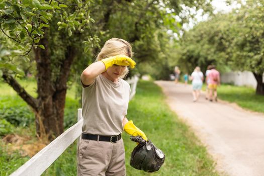 woman wearing latex gloves picking up litter to garbage bag, holding used plastic bottle in hand, takes care of planet.