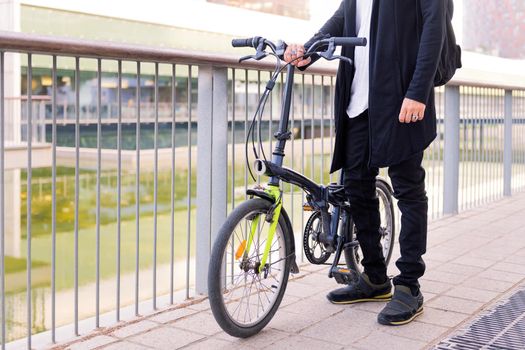 unrecognizable young man wearing trendy clothes and holding his folding bike in a city park, concept of urban lifestyle and sustainable mobility, copy space for text