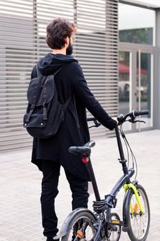 rear view of a young man wearing trendy clothes and walking with his folding bike in the city, concept of urban lifestyle and sustainable mobility