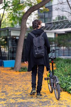 rear view of an unrecognizable college student walking with a folding bike in the park of the university campus, concept of youth and education