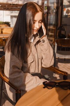 A stylish brunette woman in sunglasses is sitting at a table in an outdoor cafe with coffee and a phone. The girl is talking on the phone. Autumn walk.