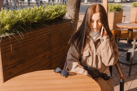 A stylish brunette woman in sunglasses is sitting at a table in an outdoor cafe with coffee and a phone. The girl is talking on the phone. Autumn walk.