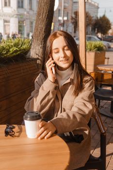 A stylish brunette woman is sitting at a table in an outdoor cafe with coffee and a phone. The girl is talking on the phone. Autumn walk.