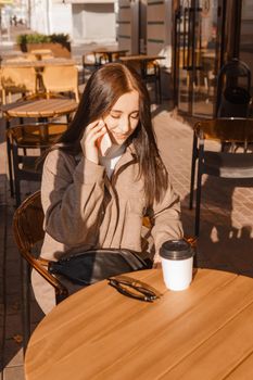 A stylish brunette woman is sitting at a table in an outdoor cafe with coffee and a phone. The girl is talking on the phone. Autumn walk.