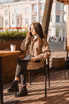 A stylish brunette woman is sitting at a table in an outdoor cafe with coffee and a phone. The girl is talking on the phone. Autumn walk.