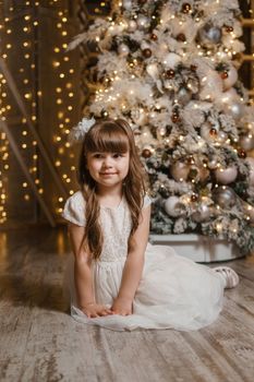 A girl in a festive light dress next to a Christmas tree, lights of garlands in the background. The concept of New Year holidays.