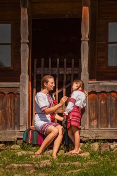 mother and daughter in Ukrainian national costumes are sitting near an old house