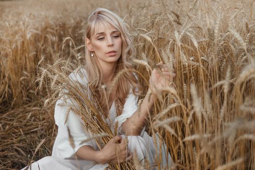 A blonde woman in a long white dress walks in a wheat field. The concept of a wedding and walking in nature.