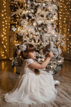 A girl in a festive light dress next to a Christmas tree, lights of garlands in the background. The concept of New Year holidays.