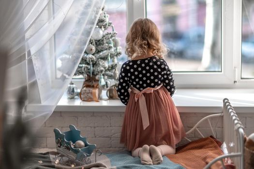 A little girl is sitting on the bed by the window and decorating a small tree with tiny Christmas toys. Happy healthy child celebrating a traditional family holiday. Adorable baby