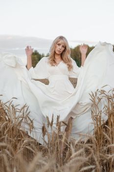 A blonde woman in a long white dress walks in a wheat field. The concept of a wedding and walking in nature.