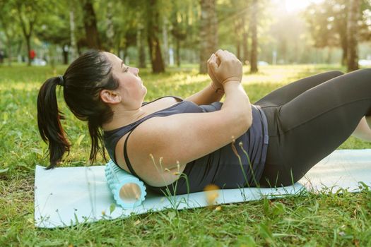 A charming brunette woman plus-size body positive practices sports in nature. Woman is engaged with a massage roller for the body in the park on a sports mat.