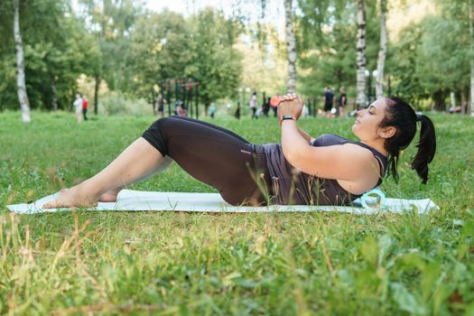 A charming brunette woman plus-size body positive practices sports in nature. Woman is engaged with a massage roller for the body in the park on a sports mat.