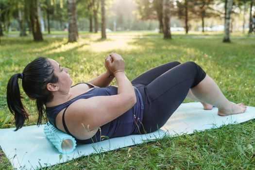 A charming brunette woman plus-size body positive practices sports in nature. Woman is engaged with a massage roller for the body in the park on a sports mat.