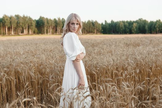 A blonde woman in a long white dress walks in a wheat field. The concept of a wedding and walking in nature.