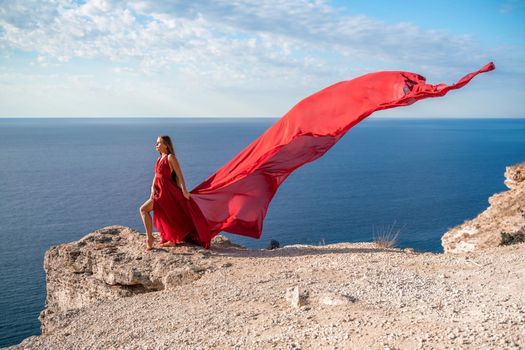 A girl with loose hair in a red dress stands on a rock rock above the sea. In the background, the sea and the rocks. The concept of travel.