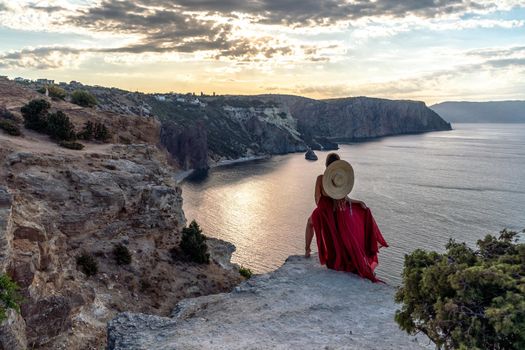 A girl with loose hair in a red dress and hat stands on the rocks above the sea. In the background, the sea and the rocks. The concept of travel