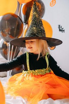 Children's Halloween - a girl in a witch hat and a carnival costume with airy orange and black balloons at home. Ready to celebrate Halloween.