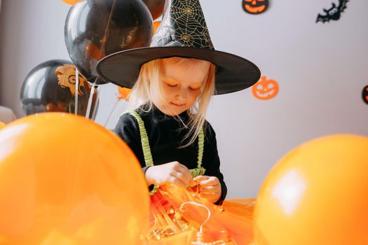 Children's Halloween - a girl in a witch hat and a carnival costume with airy orange and black balloons at home. Ready to celebrate Halloween.