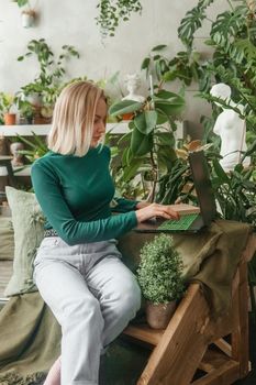 A blonde woman in a room with a lot of green indoor plants is working on a laptop. The concept of biophysical design in the interior. Work from home, work as a freelancer.