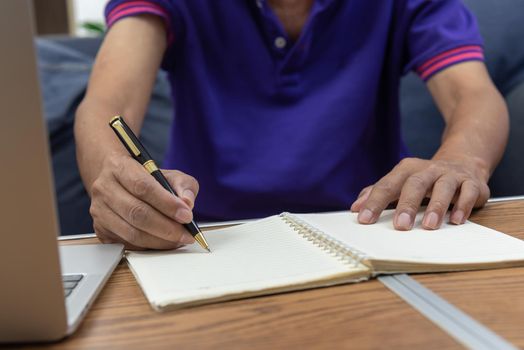 Asian elderly man holding pen write message note and computer laptop on table at sofa.