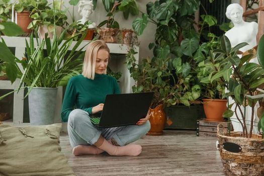 A blonde woman in a room with a lot of green indoor plants is working on a laptop. The concept of biophysical design in the interior. Work from home, work as a freelancer.