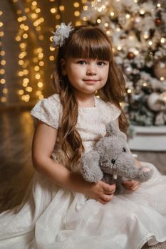 A girl in a festive light dress with a plush toy in her hands next to a Christmas tree, lights of garlands in the background. The concept of New Year holidays.