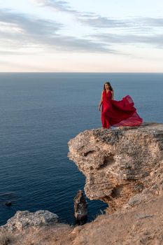 A girl with loose hair in a red dress stands on a rock rock above the sea. In the background, the sea and the rocks. The concept of travel.