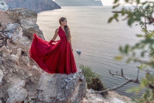 A girl with loose hair in a red dress stands on a rock rock above the sea. In the background, the sea and the rocks. The concept of travel.