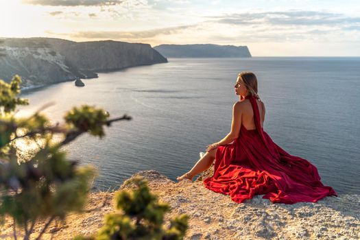 A girl with loose hair in a red dress sits on a rock rock above the sea. In the background, the sun rises from behind the mountains